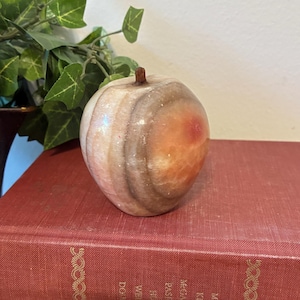 May include: A decorative apple-shaped sculpture made of stone, featuring a blend of beige, brown, and orange hues. The sculpture has a small, brown stem and sits atop a stack of vintage books with red covers. The books have gold lettering.