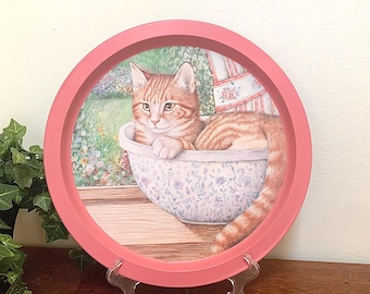 May include: A round, pink-framed decorative tray featuring a painting of a ginger cat resting in a floral bowl. The cat is lying down, with its tail curled. The background shows a garden scene. The tray is displayed on a wooden surface.