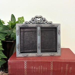 May include: A rectangular double photo frame with a metallic silver finish, featuring a decorative bow at the top and a beaded border with leaf accents. The frame has two side-by-side openings with black inserts, showcasing a textured surface.