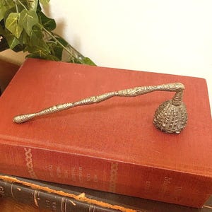 May include: A silver-toned candle snuffer with a branch-like handle and a textured, bell-shaped head rests on a vintage red book. The snuffer is positioned on top of the book, which is stacked on another antique book. Green ivy is in the background.