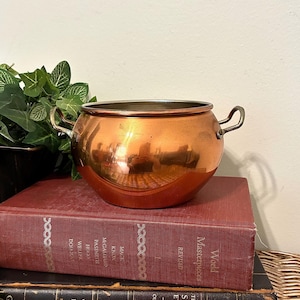 May include: A polished copper pot with a rounded body and two small metal side handles. The pot features a smooth, reflective surface and a wide circular opening at the top, showcasing a metallic interior rim and a warm, reddish-brown exterior finish.