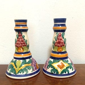 May include: Pair of ceramic candle holders. Each holder features a white base with colorful floral and geometric designs, including red, green, orange, and purple accents. The top and base are trimmed with blue and orange bands.