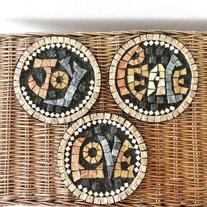Pode incluir: Três bases redondas de mosaico com as palavras "JOY", "PEACE" e "LOVE". Cada base tem um centro preto com a palavra em azulejos de mosaico coloridos. As bases estão em uma bandeja de vime tecido.