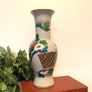 May include: A tall, off-white ceramic vase with floral and geometric patterns in red, green, blue, and white. The vase is displayed on top of two antique books with red and brown covers, and a wicker basket.