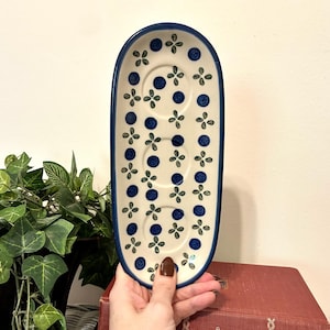 May include: An elongated oval ceramic tray with a cream-colored base, featuring a hand-painted pattern of dark blue dots and small green four-leaf floral motifs. The tray has a solid dark blue rim and three circular indentations along the center.