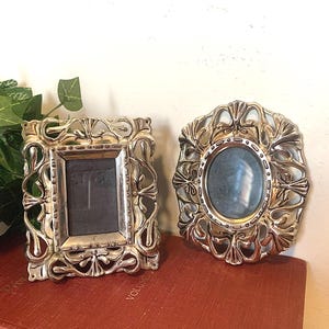 May include: Two ornate silver picture frames. One is rectangular, the other oval. Both have intricate floral and scrollwork designs. They sit on a red book, with a green plant in the background.