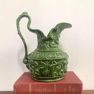 May include: A green ceramic pitcher with a curved handle and ornate relief details. The pitcher is placed on top of two antique books with red and brown covers. The pitcher's design includes figures and floral patterns.