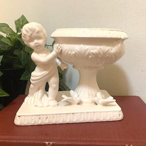May include: Off-white ceramic planter with a cherub figure. The planter has a large bowl-shaped top with leaf detailing, supported by a pedestal base. The cherub is draped in fabric and appears to be holding the planter. The base has decorative birds.