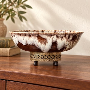 May include: A ceramic bowl featuring a drip-glaze pattern in shades of dark brown and cream. The bowl is supported by a circular, ornate gold-toned metal base with a filigree design and small, dark, rounded feet that elevate the vessel.