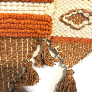 May include: Close-up of a woven wall hanging with a geometric design. The piece features a combination of orange, white, and brown yarns. Brown tassels hang from the bottom edge, adding texture and visual interest to the textile.
