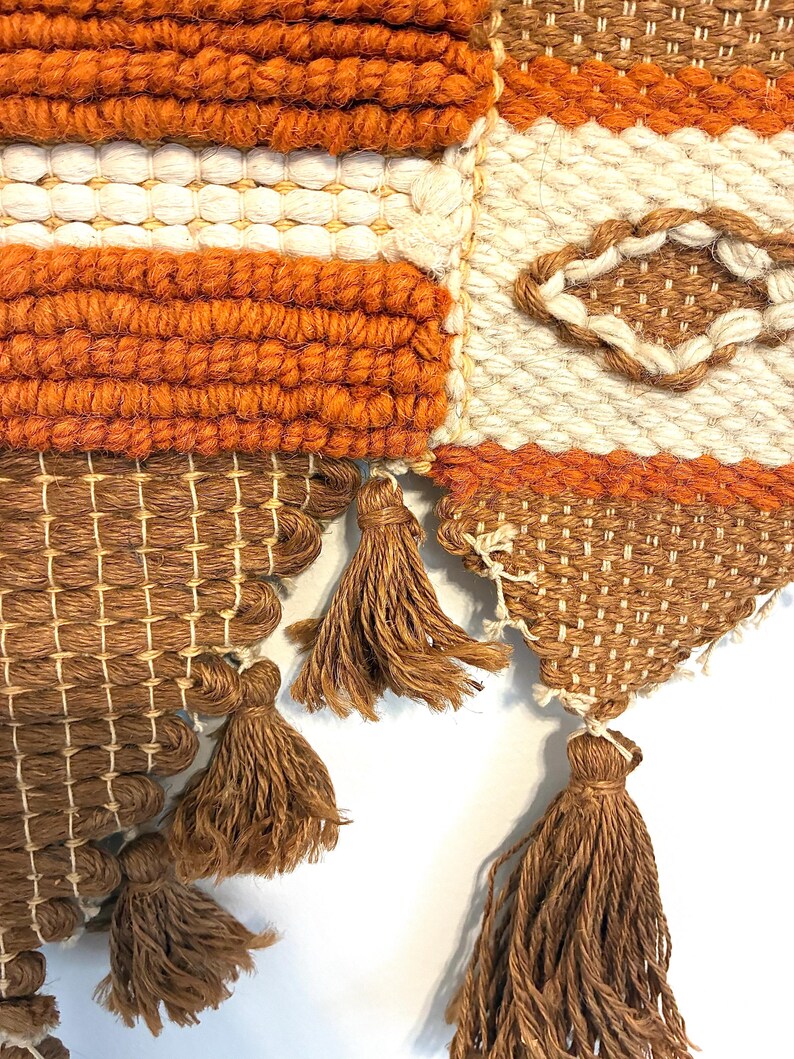 May include: Close-up of a woven wall hanging with a geometric design. The piece features a combination of orange, white, and brown yarns. Brown tassels hang from the bottom edge, adding texture and visual interest to the textile.