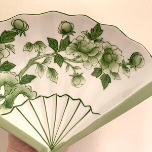 Op de afbeelding: Een wit keramisch waaiervormig bord met een groen bloemmotief. Het ontwerp toont groene bloemen, knoppen en bladeren op een tak. De rand en de lijnen van de waaier zijn ook groen.