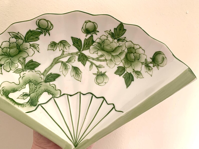 Op de afbeelding: Een wit keramisch waaiervormig bord met een groen bloemmotief. Het ontwerp toont groene bloemen, knoppen en bladeren op een tak. De rand en de lijnen van de waaier zijn ook groen.