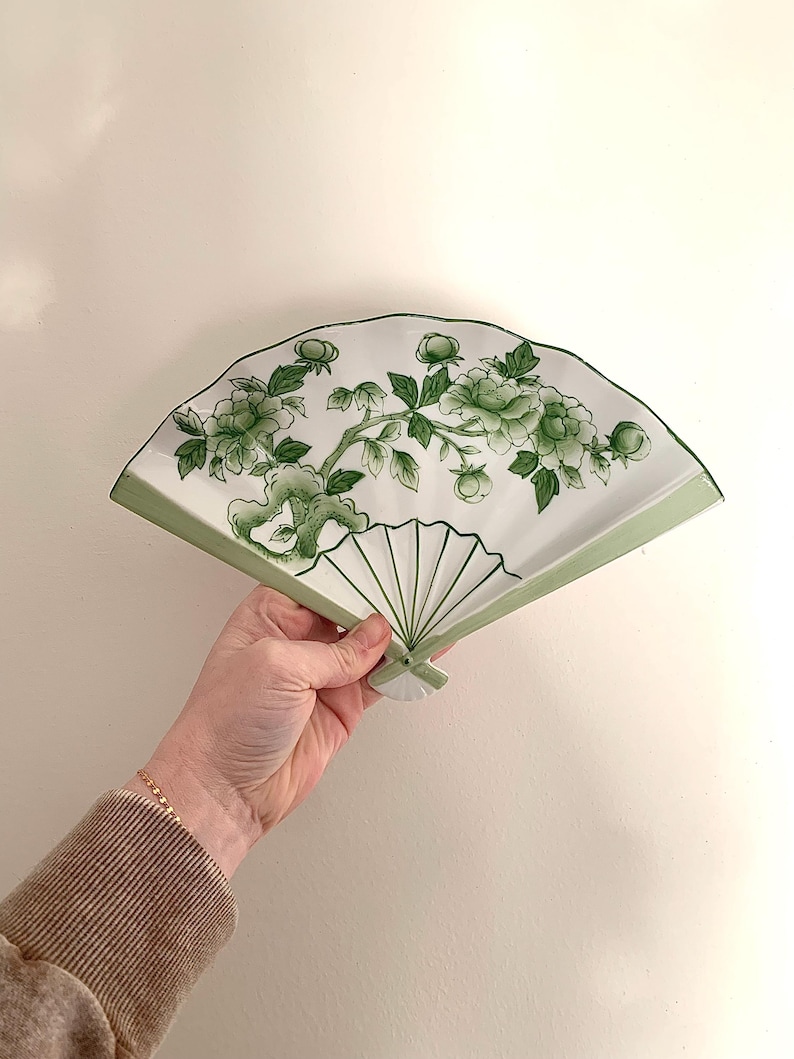 Op de afbeelding: Een decoratieve waaier in wit en groen, met een bloemenontwerp. De waaier heeft een groen bloemenpatroon op een witte achtergrond, met een groene rand. De waaier wordt tegen een witte muur gehouden.