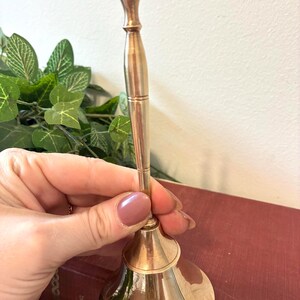 Op de afbeelding: Een messingkleurige handbel met een lange handgreep en een uitlopende basis. De bel wordt in een hand gehouden, met een vage achtergrond van groen en een rood boek. De bel wordt waarschijnlijk gebruikt om te signaleren of te decoreren.