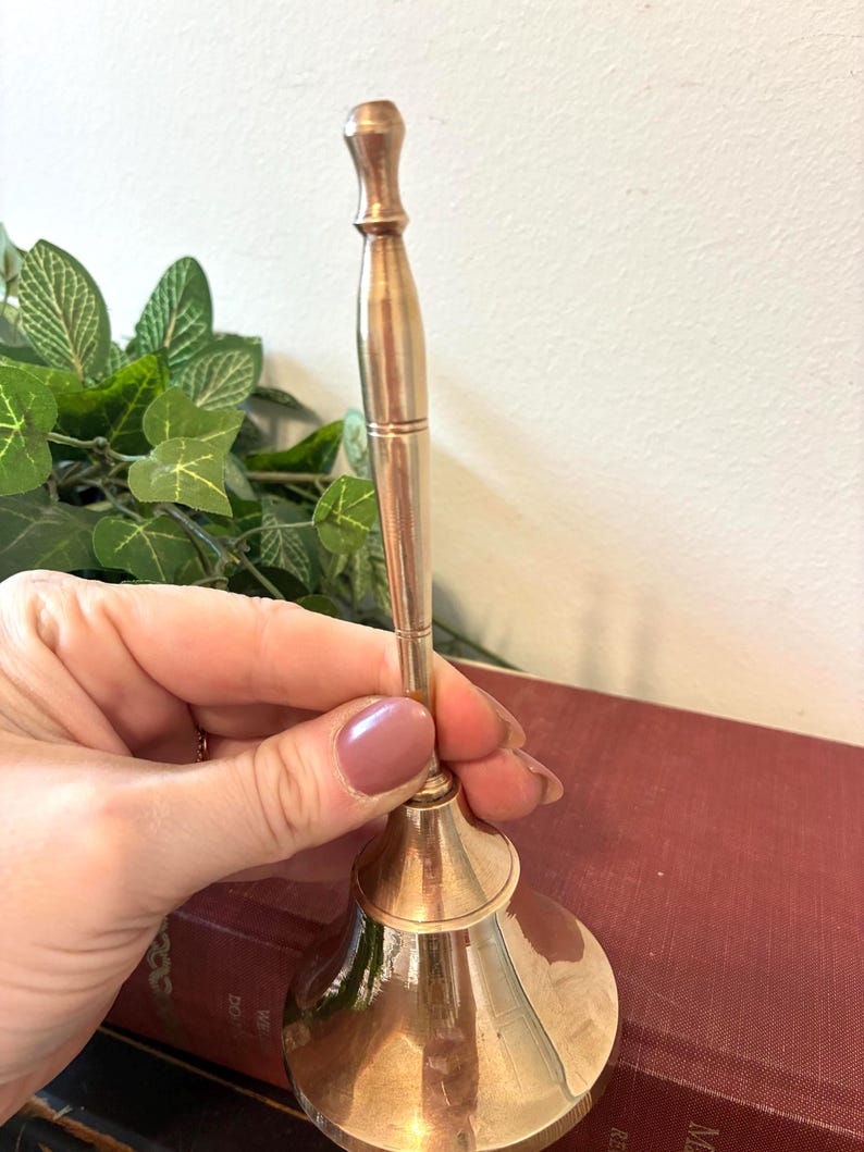 Op de afbeelding: Een messingkleurige handbel met een lange handgreep en een uitlopende basis. De bel wordt in een hand gehouden, met een vage achtergrond van groen en een rood boek. De bel wordt waarschijnlijk gebruikt om te signaleren of te decoreren.