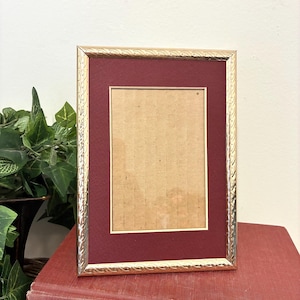 May include: A rectangular picture frame with a metallic gold finish featuring a textured, braided pattern along the border. The frame includes a deep burgundy matting surrounding a central rectangular opening with a light tan cardboard backing.