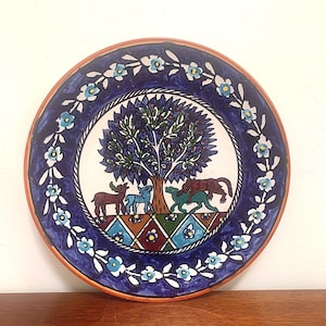 May include: A round, hand-painted ceramic plate with a vibrant design. The plate features a central image of a tree with animals, surrounded by a blue border with floral patterns. The rim is a reddish-brown color.