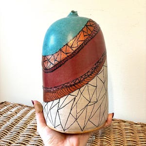 May include: A handcrafted ceramic vase with a rounded shape. The top is teal, transitioning to a maroon midsection with orange and black geometric designs. The bottom half features a cream color with black geometric patterns.