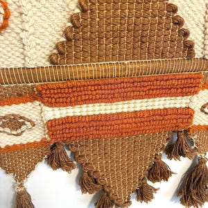 May include: A woven wall hanging with a geometric design. The piece features a combination of cream, brown, and orange yarns. The design includes diamond shapes, horizontal stripes, and tassels at the bottom. The texture is visible, showcasing the craftsmanship.