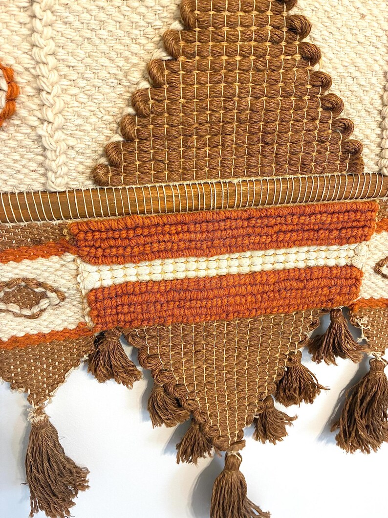 May include: A woven wall hanging with a geometric design. The piece features a combination of cream, brown, and orange yarns. The design includes diamond shapes, horizontal stripes, and tassels at the bottom. The texture is visible, showcasing the craftsmanship.