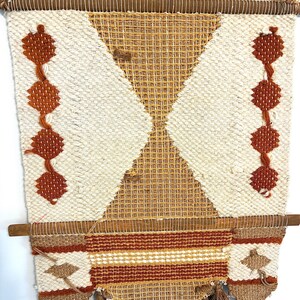 May include: Handwoven wall hanging with a geometric design. The tapestry features a cream-colored background with diamond and triangle patterns in shades of brown and tan. It includes tassels at the bottom and is suspended from a wooden rod.