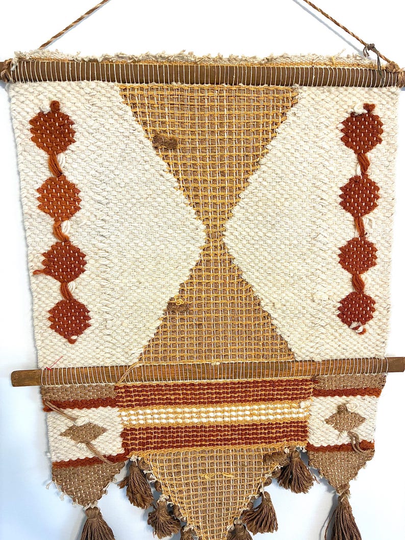 May include: Handwoven wall hanging with a geometric design. The tapestry features a cream-colored background with diamond and triangle patterns in shades of brown and tan. It includes tassels at the bottom and is suspended from a wooden rod.