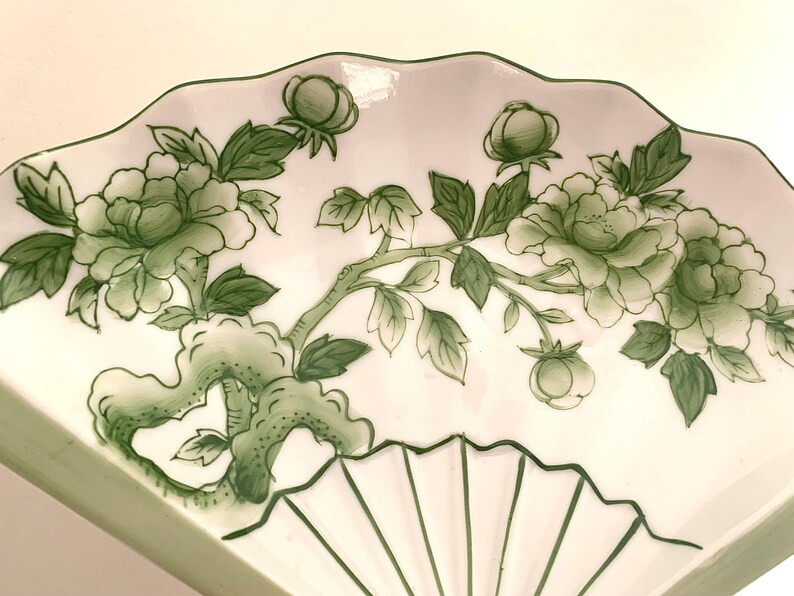 Op de afbeelding: Witte, waaiervormige decoratieve schaal met een groen bloemmotief. Het ontwerp toont groene pioenrozen, knoppen en bladeren op een tak. De schaal heeft een geschulpte rand en een waaiervormig patroon.