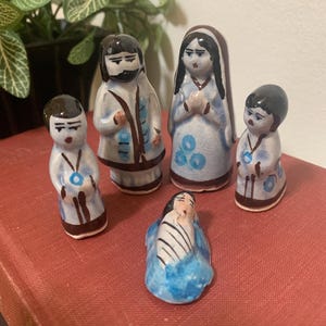 May include: A set of five ceramic figurines depicting a religious scene. The figures are painted with brown, blue, and white details. The figurines are arranged on a red surface, with a plant in the background.