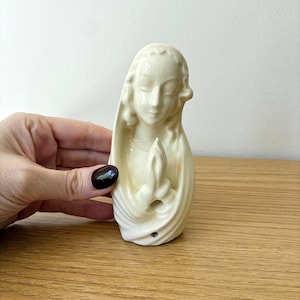 May include: A cream-colored ceramic figurine depicting a figure with a draped head covering and hands pressed together in a prayer position. The sculpture features a smooth, glossy finish and a sculpted face with closed eyes and serene features.