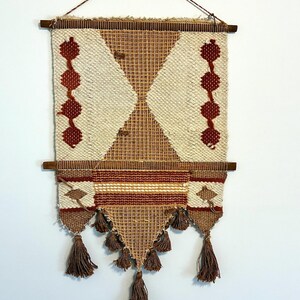 May include: A woven wall hanging with a geometric design. The piece features a cream-colored background with brown and red accents. It has a diamond pattern in the center, with decorative tassels at the bottom and is suspended from a wooden rod.