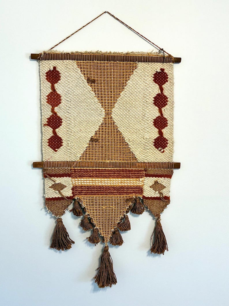 May include: A woven wall hanging with a geometric design. The piece features a cream-colored background with brown and red accents. It has a diamond pattern in the center, with decorative tassels at the bottom and is suspended from a wooden rod.