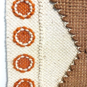 May include: A woven wall hanging with a geometric design. The piece features a cream-colored background with three orange and white circular accents. A brown woven section forms a diagonal pattern. The piece is mounted on a wooden dowel.