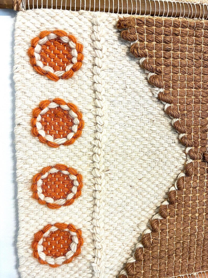 May include: A woven wall hanging with a geometric design. The piece features a cream-colored background with three orange and white circular accents. A brown woven section forms a diagonal pattern. The piece is mounted on a wooden dowel.