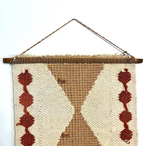 May include: Woven wall hanging with a geometric design. The piece features a cream-colored background with a diamond-shaped pattern in tan and rust-colored accents. Two vertical lines of rust-colored diamond shapes are on either side. The piece is mounted on a wooden dowel.