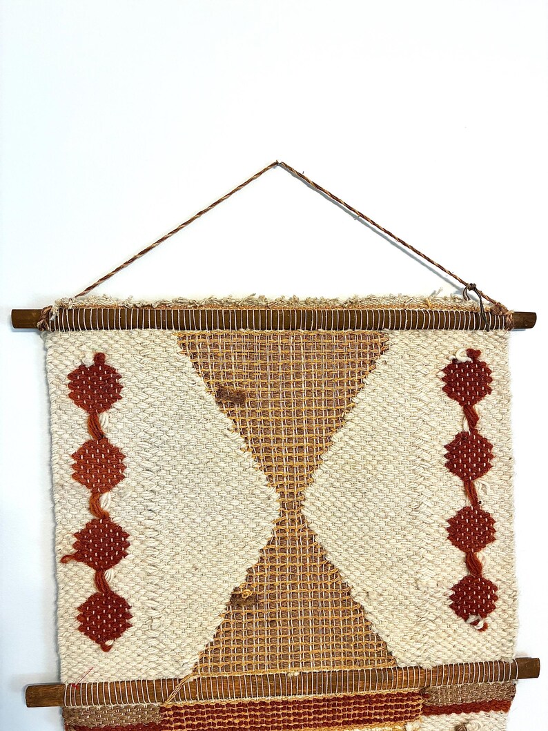 May include: Woven wall hanging with a geometric design. The piece features a cream-colored background with a diamond-shaped pattern in tan and rust-colored accents. Two vertical lines of rust-colored diamond shapes are on either side. The piece is mounted on a wooden dowel.