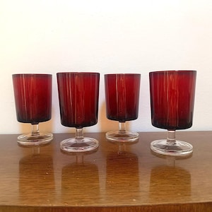May include: Four ruby red stemmed glasses with vertical ribbed detailing. The glasses have a clear base and are displayed on a wooden surface. The glasses are likely for serving beverages.