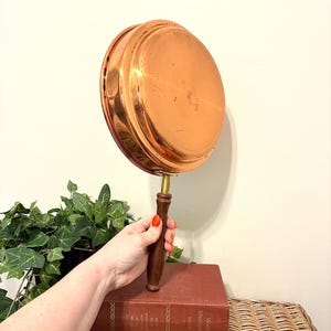 May include: A round copper pan with a polished metallic finish and a smooth, curved surface. The pan features a long, cylindrical dark brown wooden handle attached to the side with a brass-colored metal connector, showing a reflective sheen.