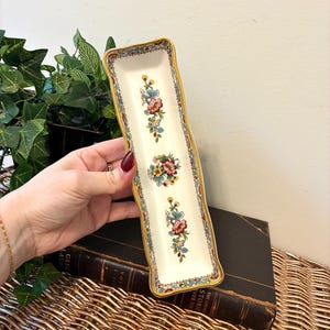 May include: A rectangular, cream-colored ceramic dish with a floral pattern. The dish has a gold trim and features three clusters of flowers in red, blue, and yellow. The dish is held in a person's hand.