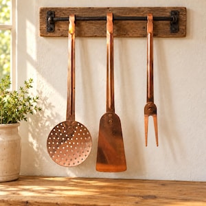 May include: A set of three copper kitchen utensils including a skimmer, a spatula, and a two-pronged fork, all hanging from a dark metal rod mounted on a rectangular piece of weathered, light brown wood attached to a wall.