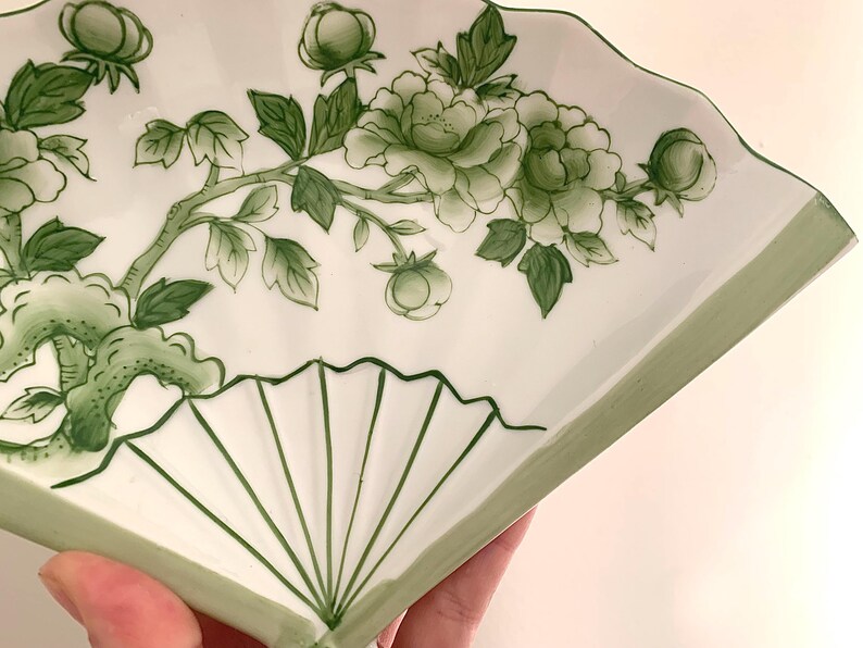 Op de afbeelding: Een wit keramisch waaiervormig bord met een groen bloemenontwerp. Het ontwerp toont groene bloemen, bladeren en knoppen op een tak. De rand en de lijnen van de waaier zijn ook groen.