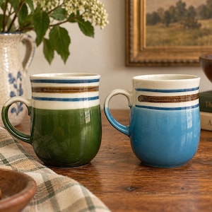 May include: Two ceramic mugs with rounded bodies and handles. One mug features a deep green base, while the other has a light blue base. Both mugs are decorated with horizontal blue and brown stripes around the upper rim and a white interior.