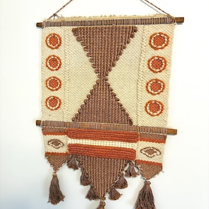 May include: A woven wall hanging with a geometric design. The piece features a cream-colored background with brown and orange accents. The design includes a central diamond shape, circular patterns, and tassels at the bottom. The wall hanging is suspended from a wooden rod.
