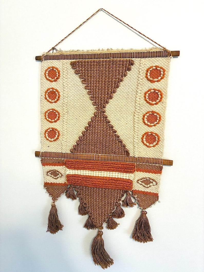 May include: A woven wall hanging with a geometric design. The piece features a cream-colored background with brown and orange accents. The design includes a central diamond shape, circular patterns, and tassels at the bottom. The wall hanging is suspended from a wooden rod.