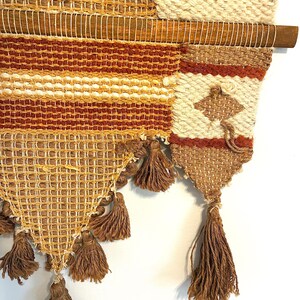 May include: A woven wall hanging with a wooden dowel. The textile features a mix of textures and colors, including beige, brown, and orange. The design incorporates geometric patterns and tassels, creating a bohemian aesthetic. The wall hanging is a decorative piece.