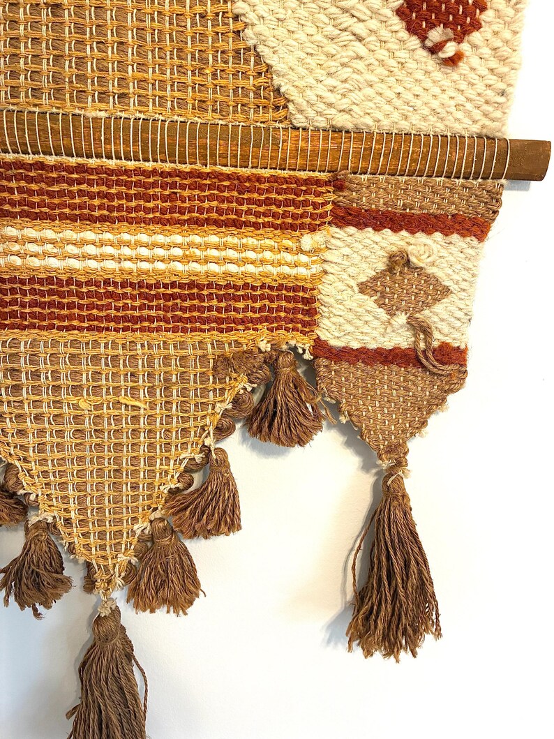May include: A woven wall hanging with a wooden dowel. The textile features a mix of textures and colors, including beige, brown, and orange. The design incorporates geometric patterns and tassels, creating a bohemian aesthetic. The wall hanging is a decorative piece.
