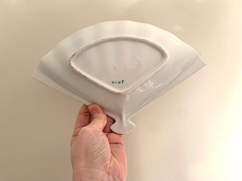 Op de afbeelding: Een witte, waaiervormige keramische schaal met een geschulpte rand. De onderkant heeft een rechthoekige basis en een blauw nummer 7. De schaal wordt door een hand vastgehouden, wat de vorm en het ontwerp laat zien.