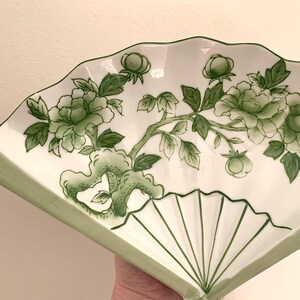 Op de afbeelding: Wit keramisch waaiervormig serveerbord met een groen bloemmotief. De schaal heeft een geschulpte rand en een geschilderde groene rand. Het bloemmotief omvat groene bladeren, knoppen en bloemen.