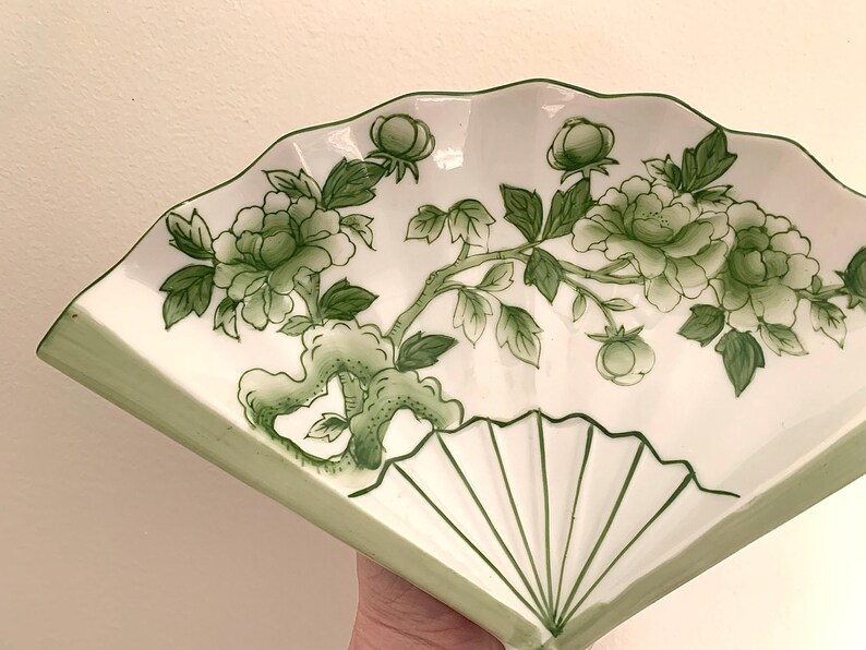 Op de afbeelding: Wit keramisch waaiervormig serveerbord met een groen bloemmotief. De schaal heeft een geschulpte rand en een geschilderde groene rand. Het bloemmotief omvat groene bladeren, knoppen en bloemen.