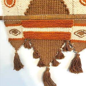 May include: Woven wall hanging with a geometric design in cream, brown, and orange. The textile features a variety of textures, including woven sections, tassels, and embroidered details. The wall hanging is mounted on a wooden dowel.